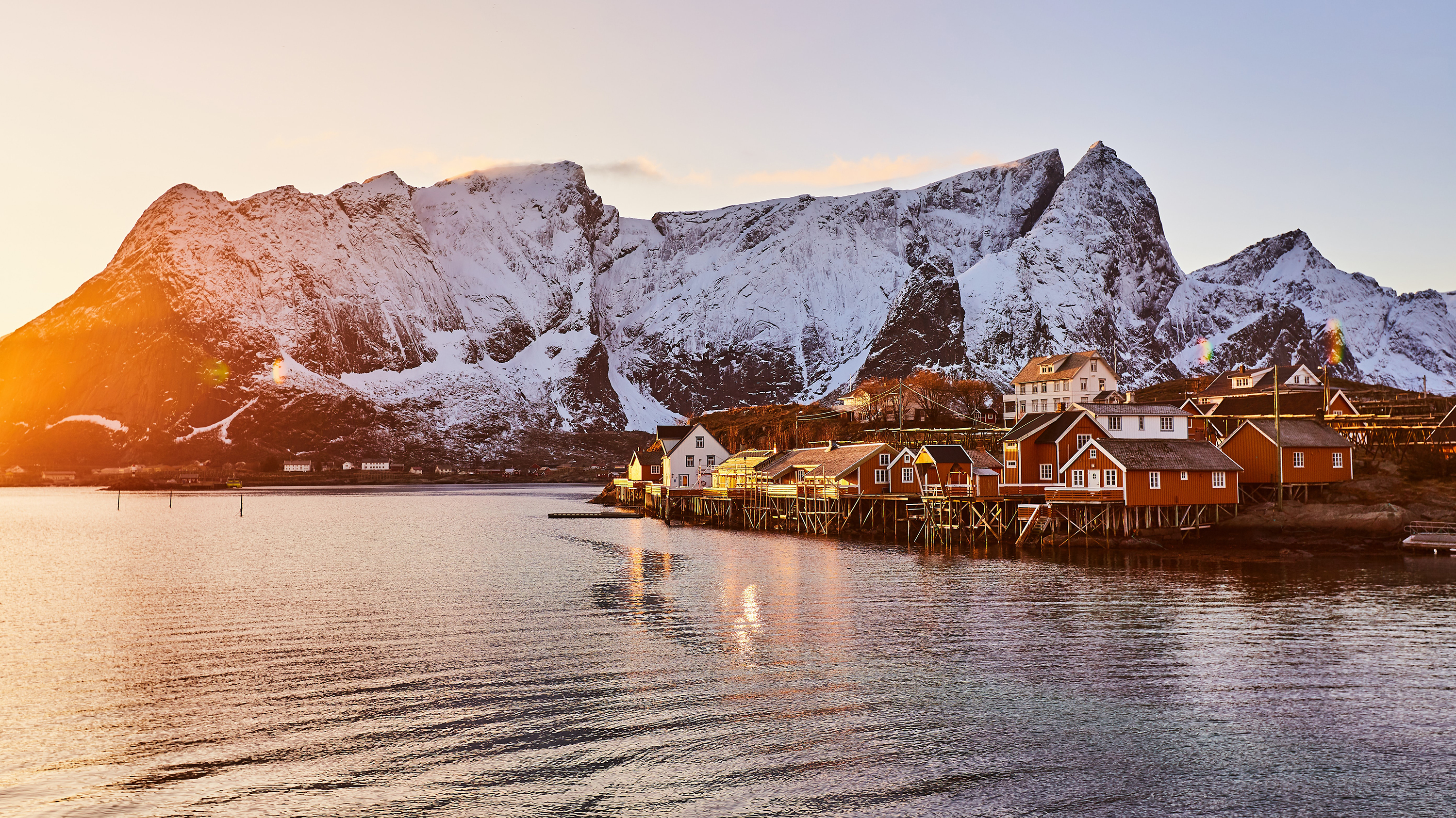 Rorbuer og hus langs sjøen i Lofoten med snødekte fjell i bakgrunnen.