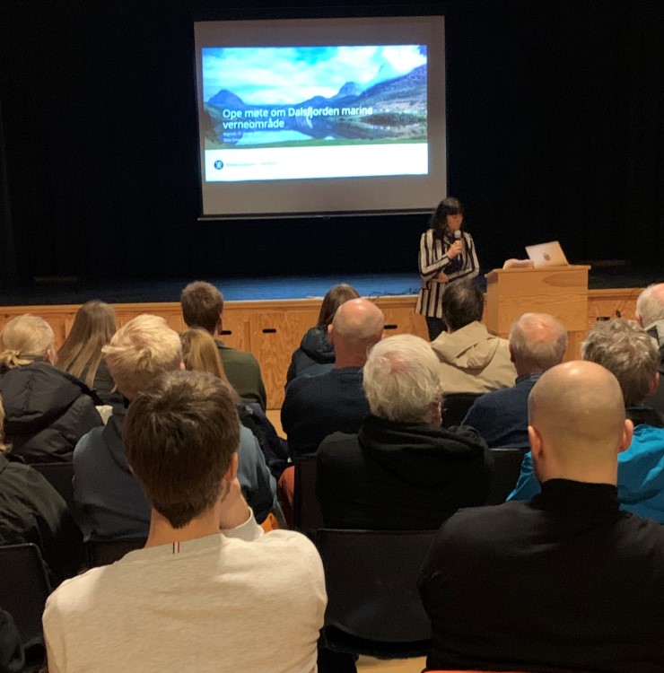 Mange hovud ser mot ein person som snakkar i mikrofon framfor ein skjerm