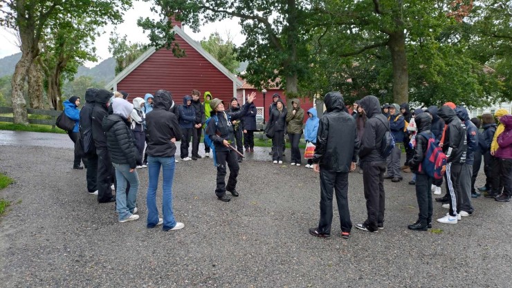 Mange menneskje står framfor store tre og ei dame peikar opp på trea. Det er vått. 