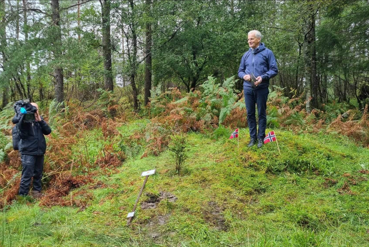 en person står i skogen og smiler og held tale på bakken er det norske flagg
