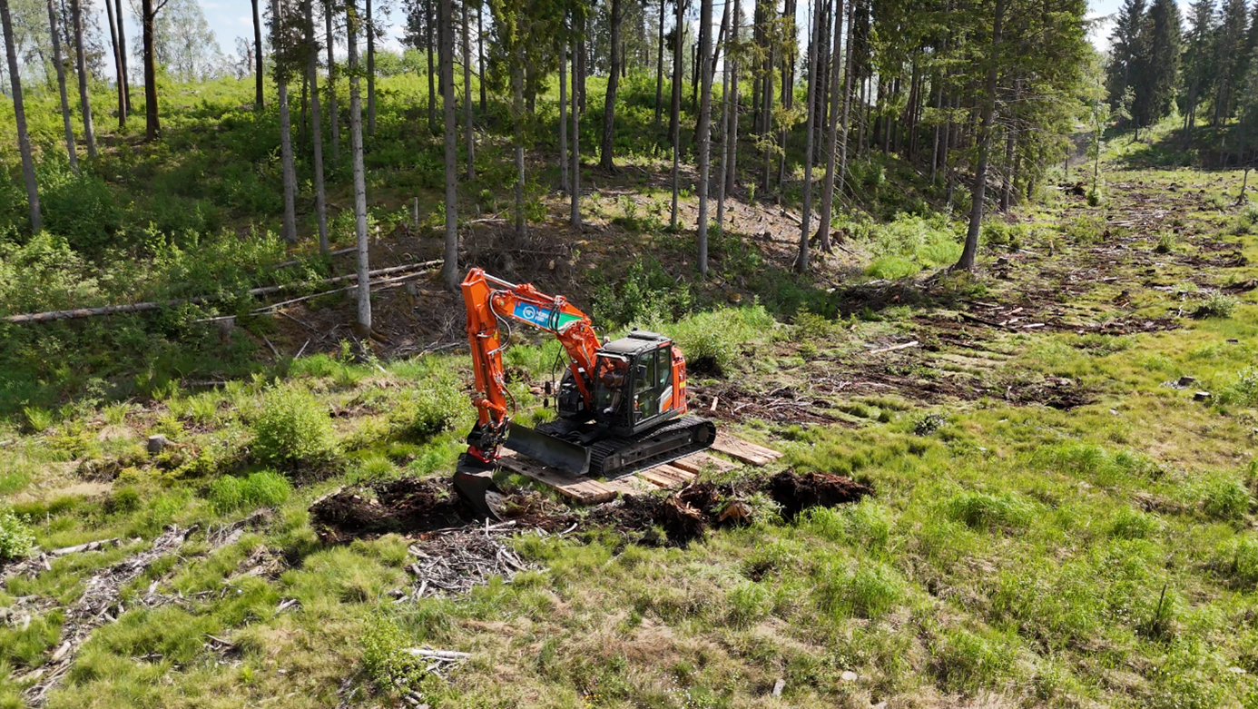 Et bilde som viser en gravemaskin på myra, grønn vegetasjon og skog. 