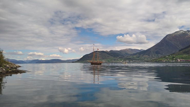 seglbåt utan segl, på fjord omkransa av høge fjell. Mindre båtar i følgje. 