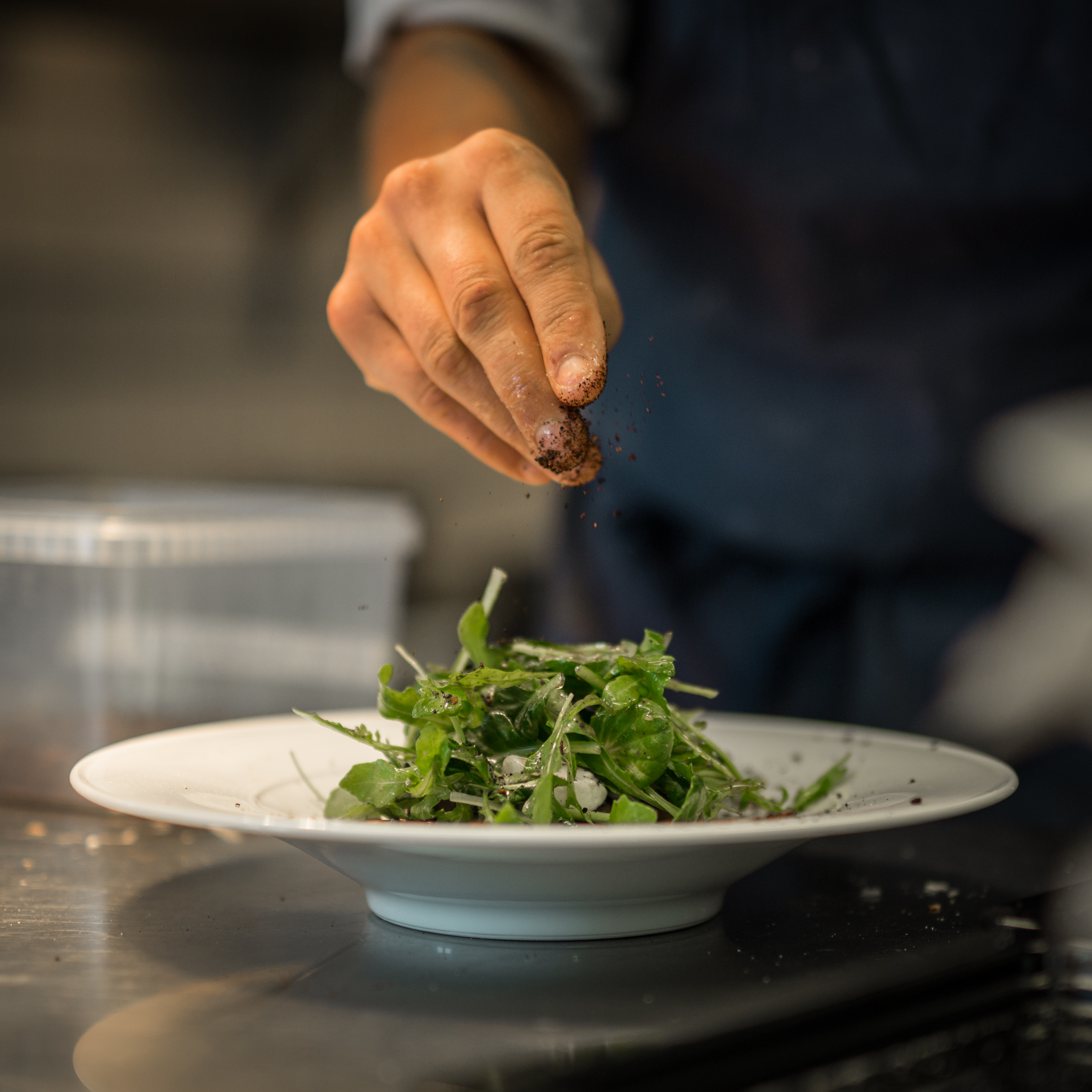 Tallerken med grønn ruccola og en hånd som strør på krydder.