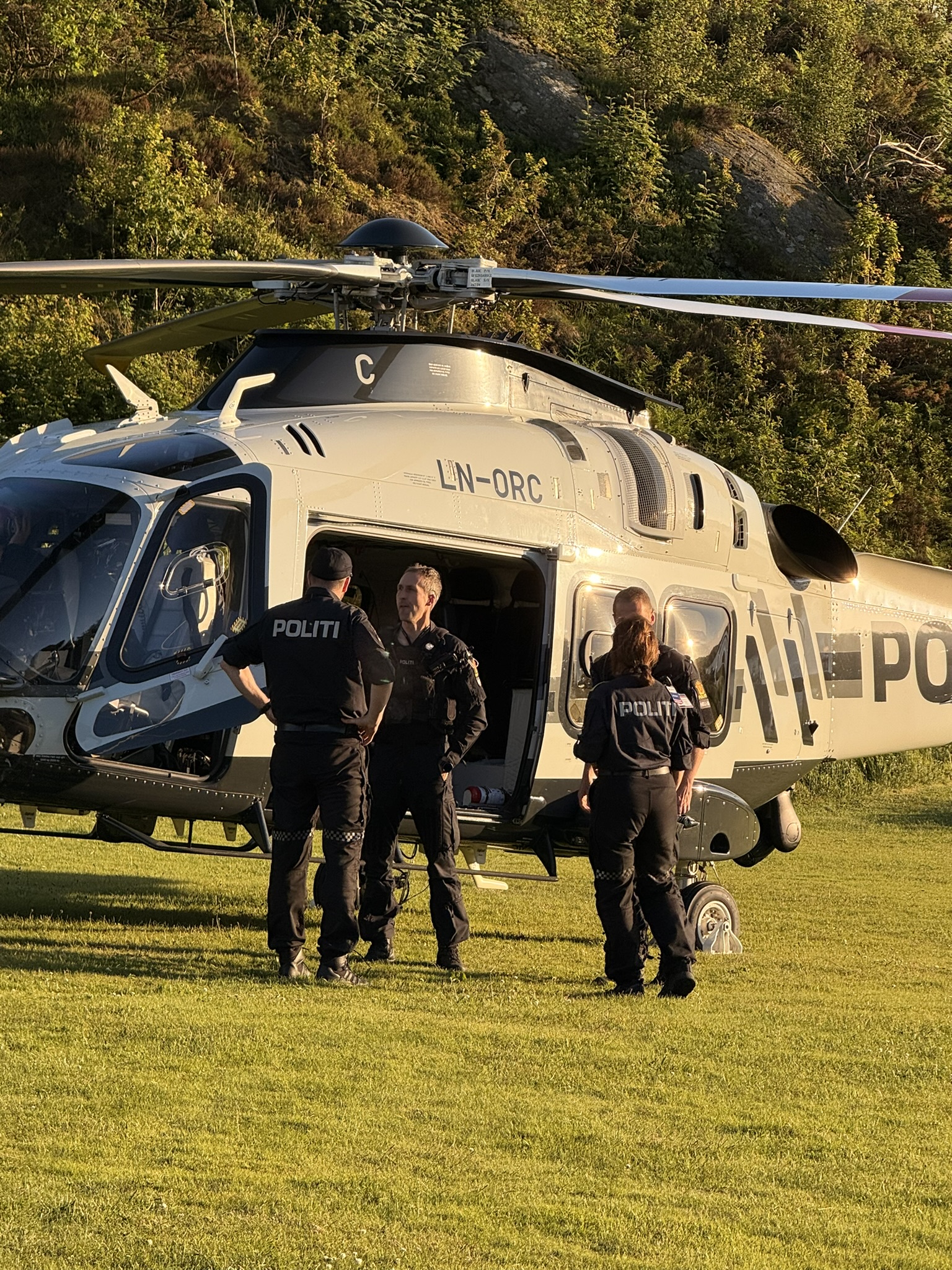 Politihelikopter parkert på et gressområde omgitt av trær, med flere uniformerte politifolk samlet ved den åpne sidedøren i varmt kveldslys.