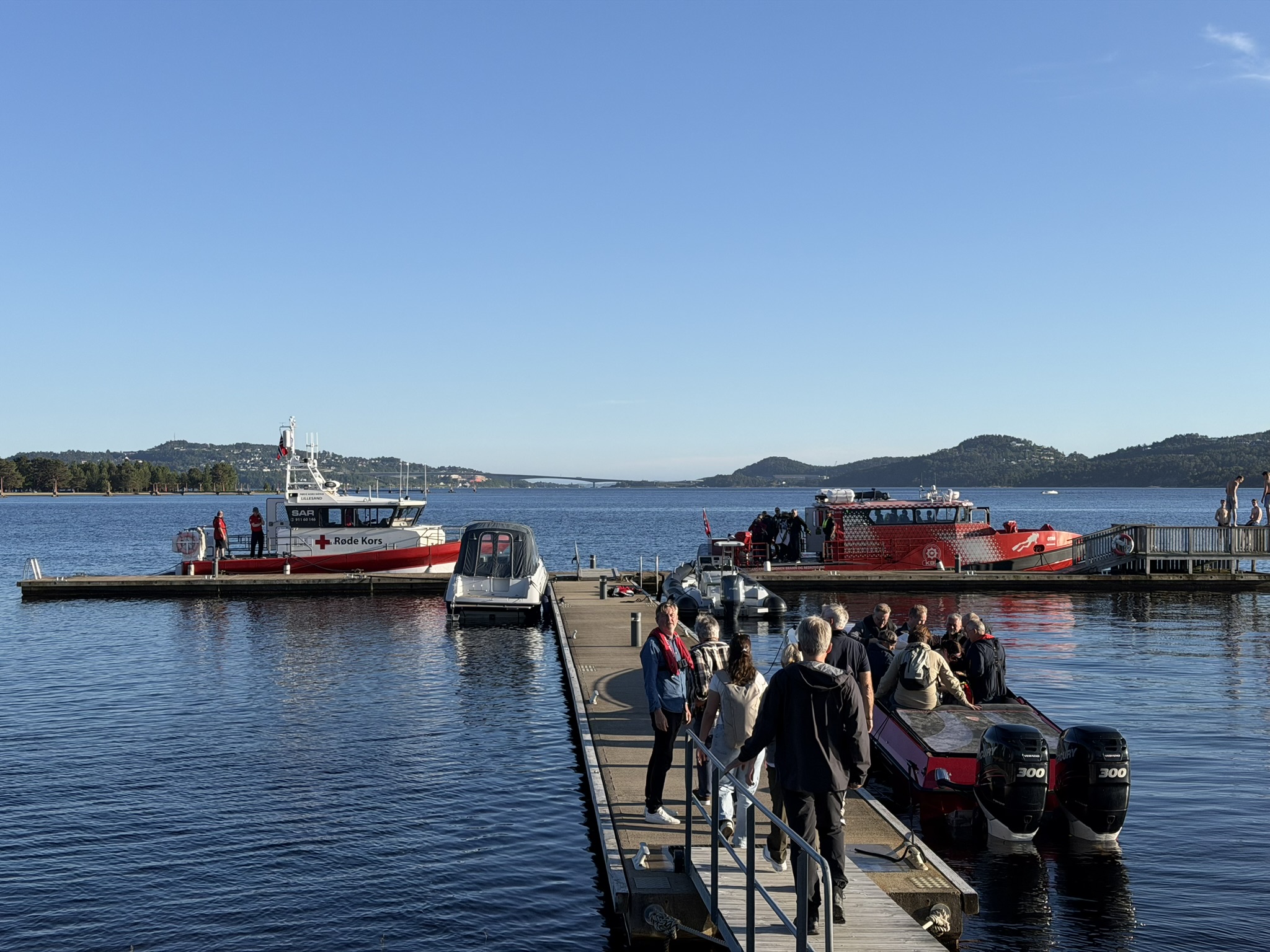 Brygge ved sjøen med flere personer som går om bord i småbåter, mens redningsbåt fra Røde Kors og en annen beredskapsbåt ligger fortøyd i rolig vann, med skjærgård og bro i bakgrunnen under klar blå himmel.