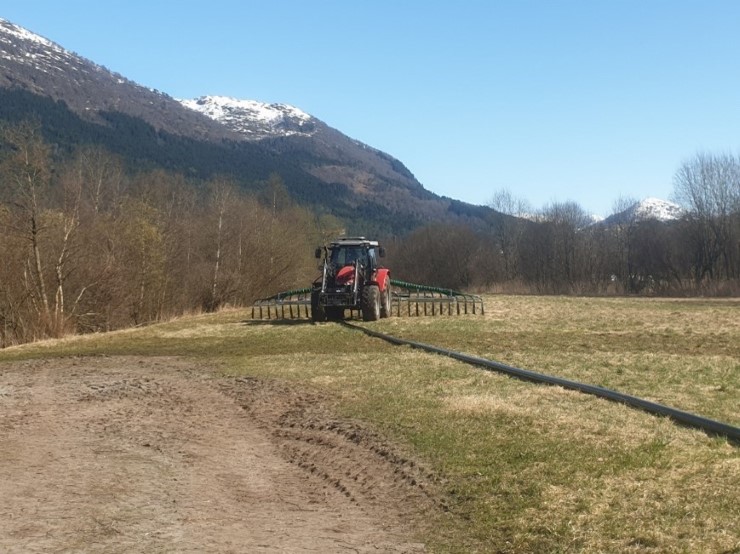 Blå himmel, fjell med snø på toppane, skog, rød traktor, nedlegging av gjødsel med nedleggingsutstyr
