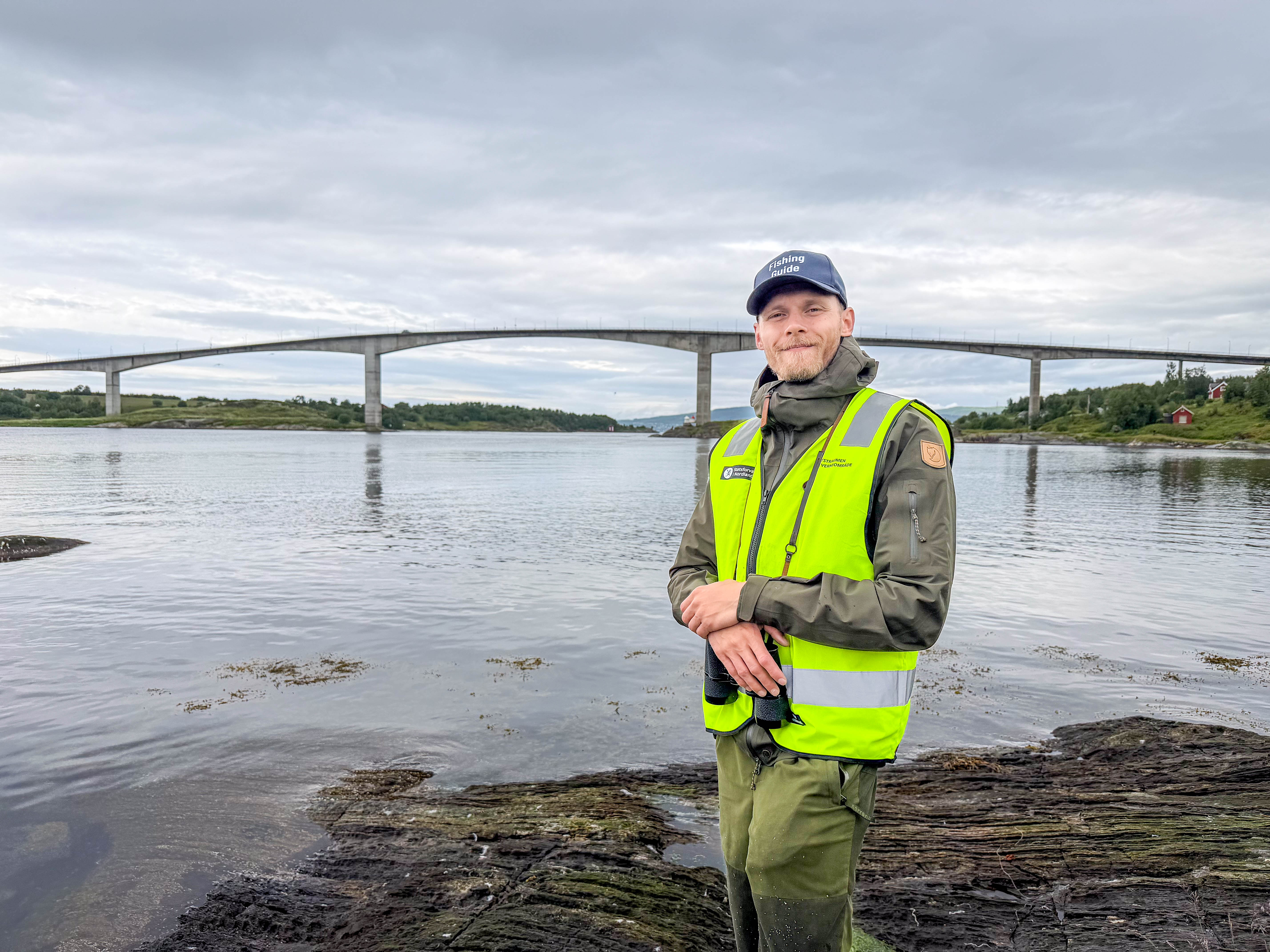 En mann med refleksvest ser i kameraet. I bakgrunn er det hav og en bru. 