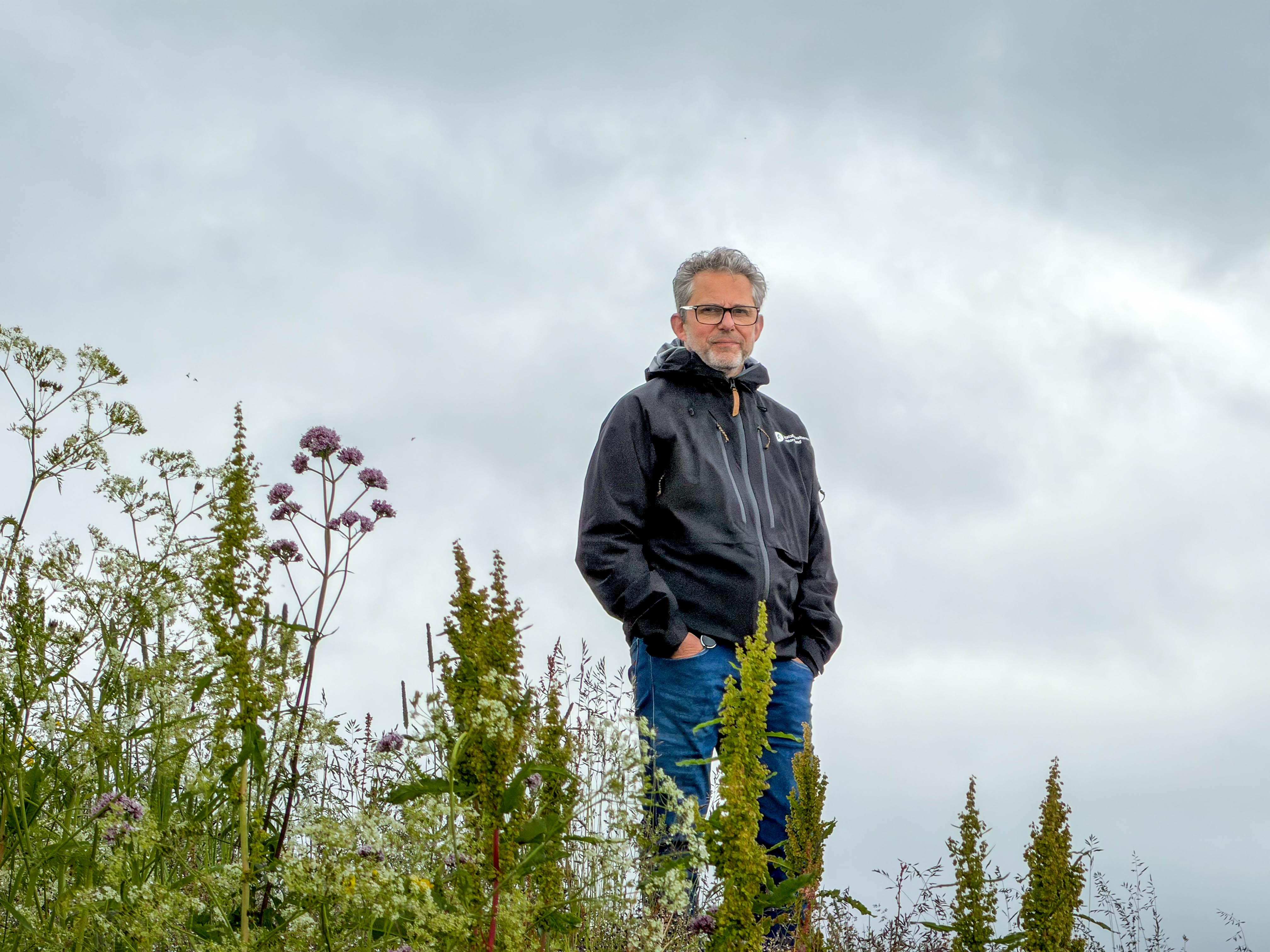 En mann står med hendene i lomma med høyt gress i forgrunnen. 