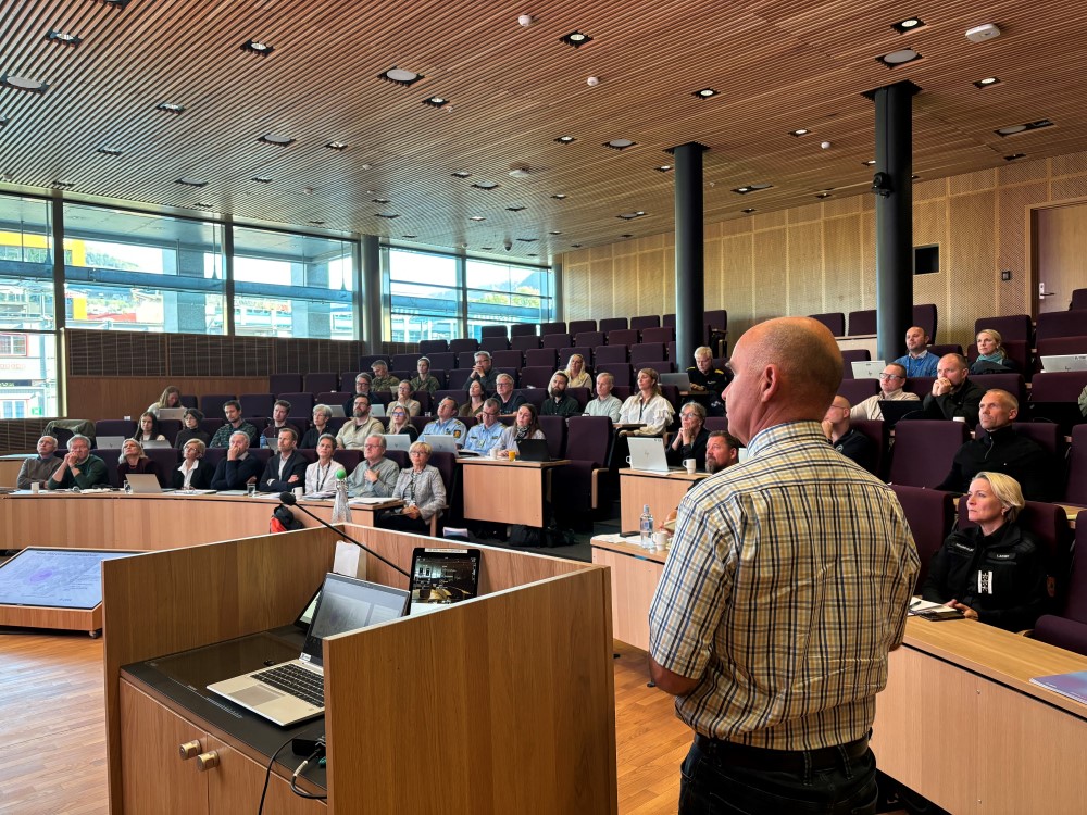 Mann ved talerstol. Mennesker i auditorium.