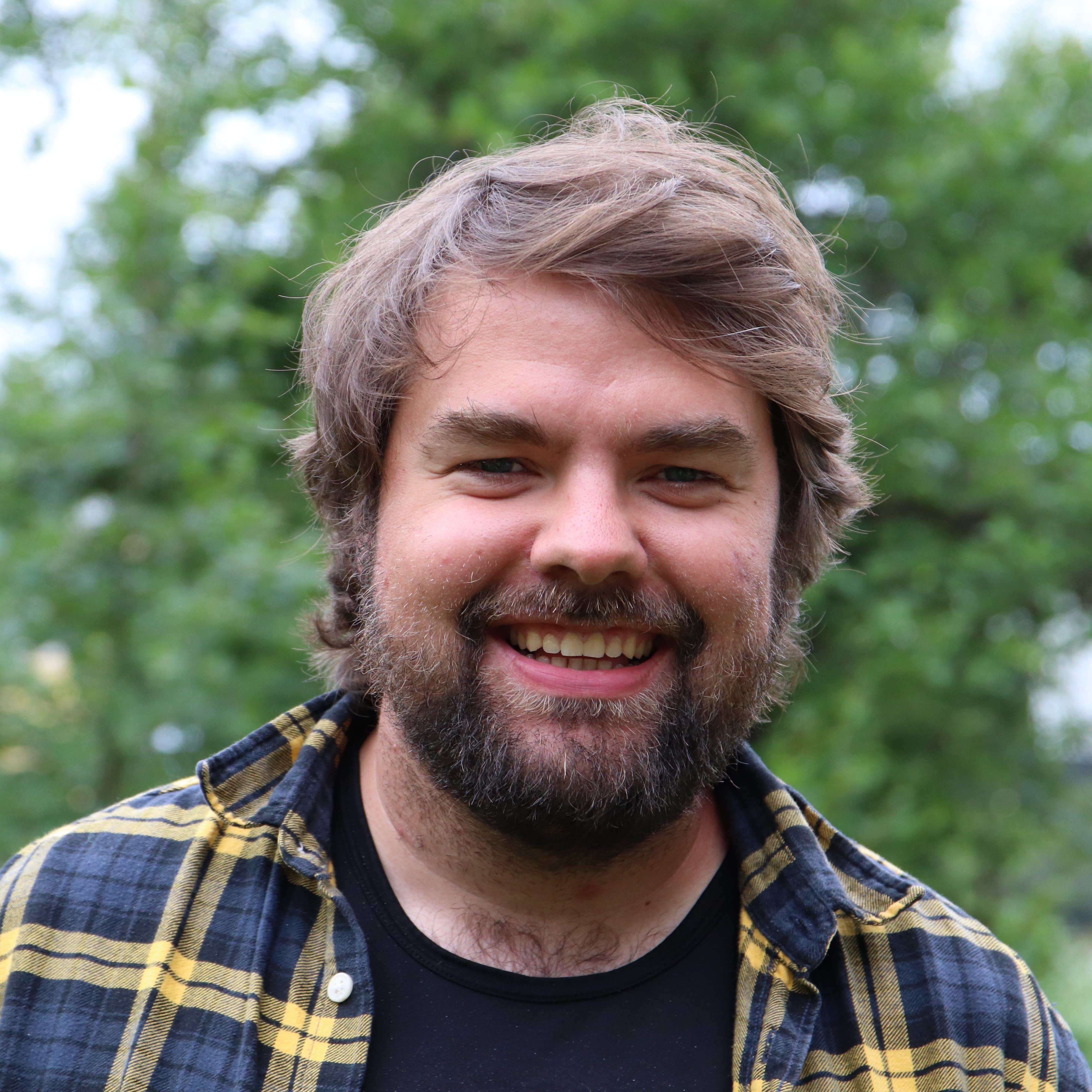 Smilende mann med mørkt kort hår og rutete skjorte. Bildet er tatt utendørs på sommeren og bakgrunnen er grønn. 