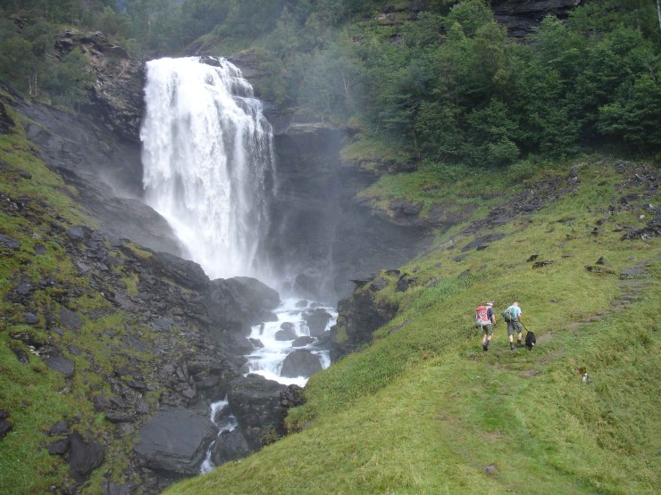 Hvit foss ned langs fjellside med frisk grønn vegetasjon langs sidene. To personer og en hund går oppover på høyre side av bildet.