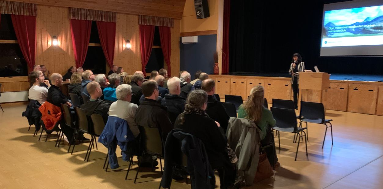 ein stor sal med folk som sit på stoler og ein dame står framfor ei skjerm som syner bilete av Dalsfjorden