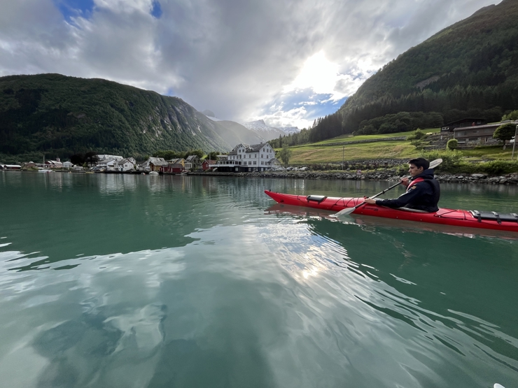 ein mann i ein kajakk langs fjord med bygd og fjell i bakgrunnen