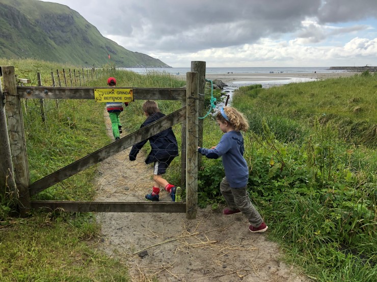 Tre barn løper gjennom ei grind for å gå til ein strand i framgrunnen