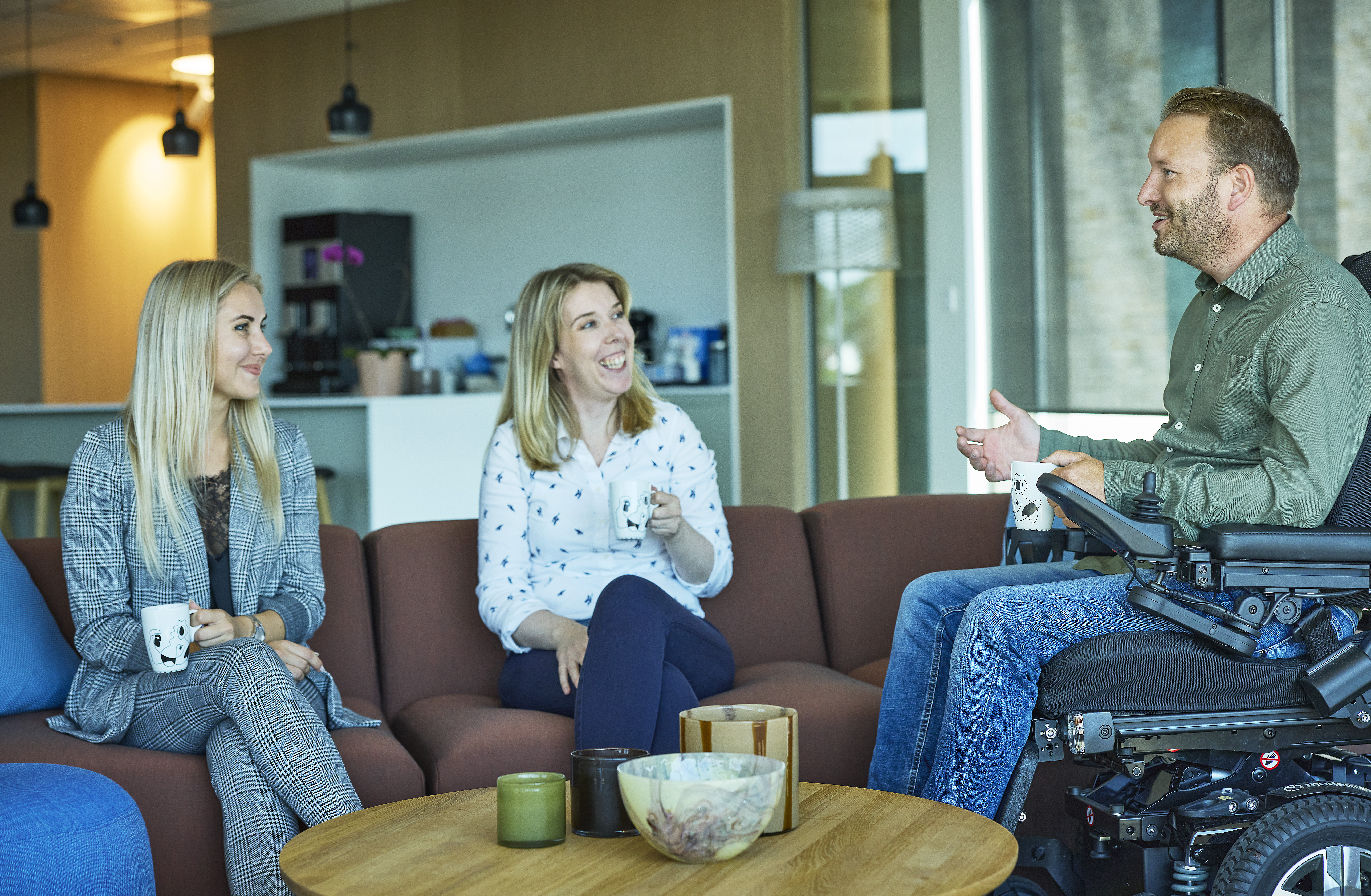 To kvinner i sofagruppe og én mann i rullestol har en samtale og ler