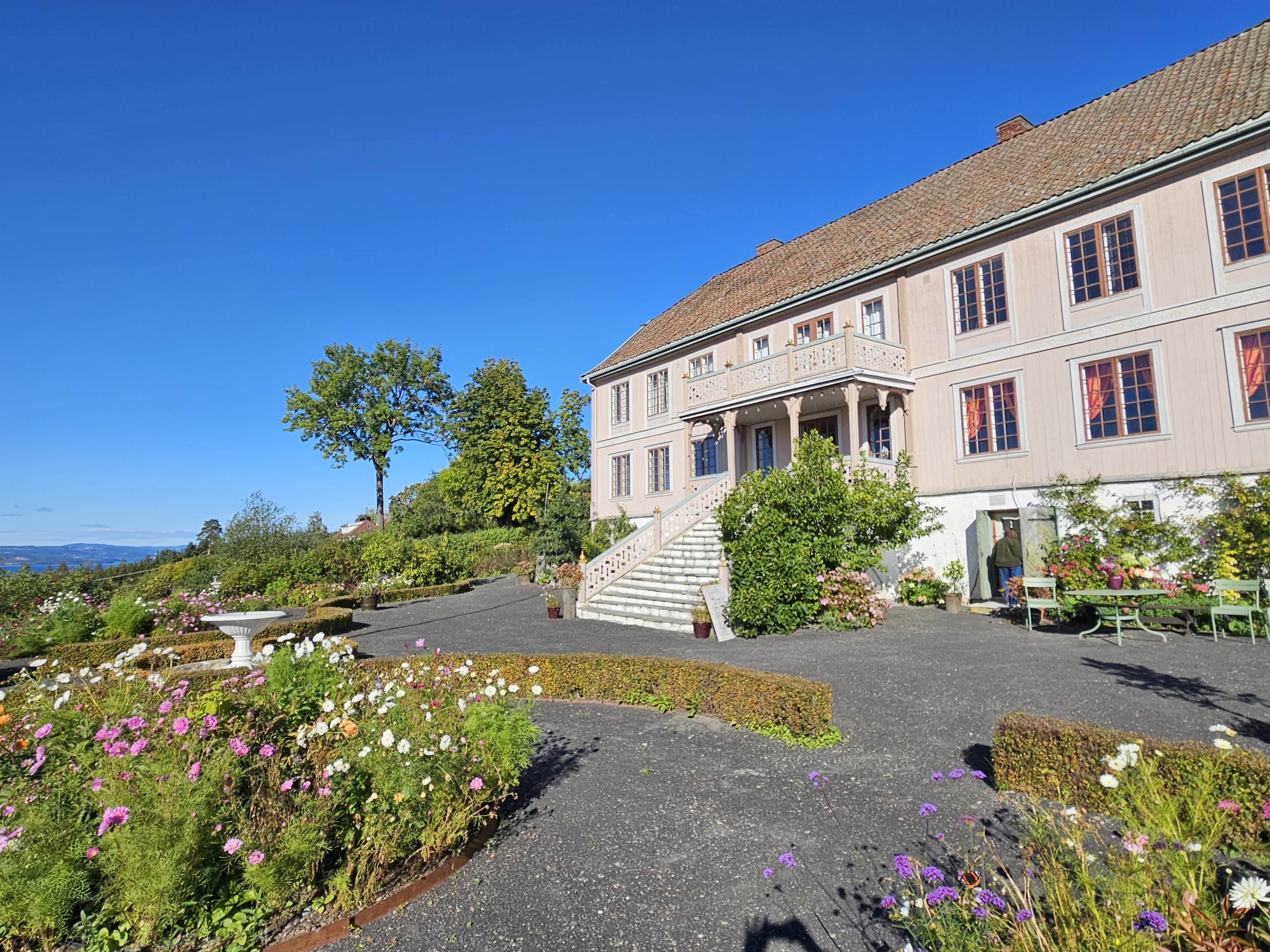 Hageanlegg med mørke grusganger og frodige blomsterbed foran en stor, blekrosa hovedbygning med brei trapp ned i hagen.
