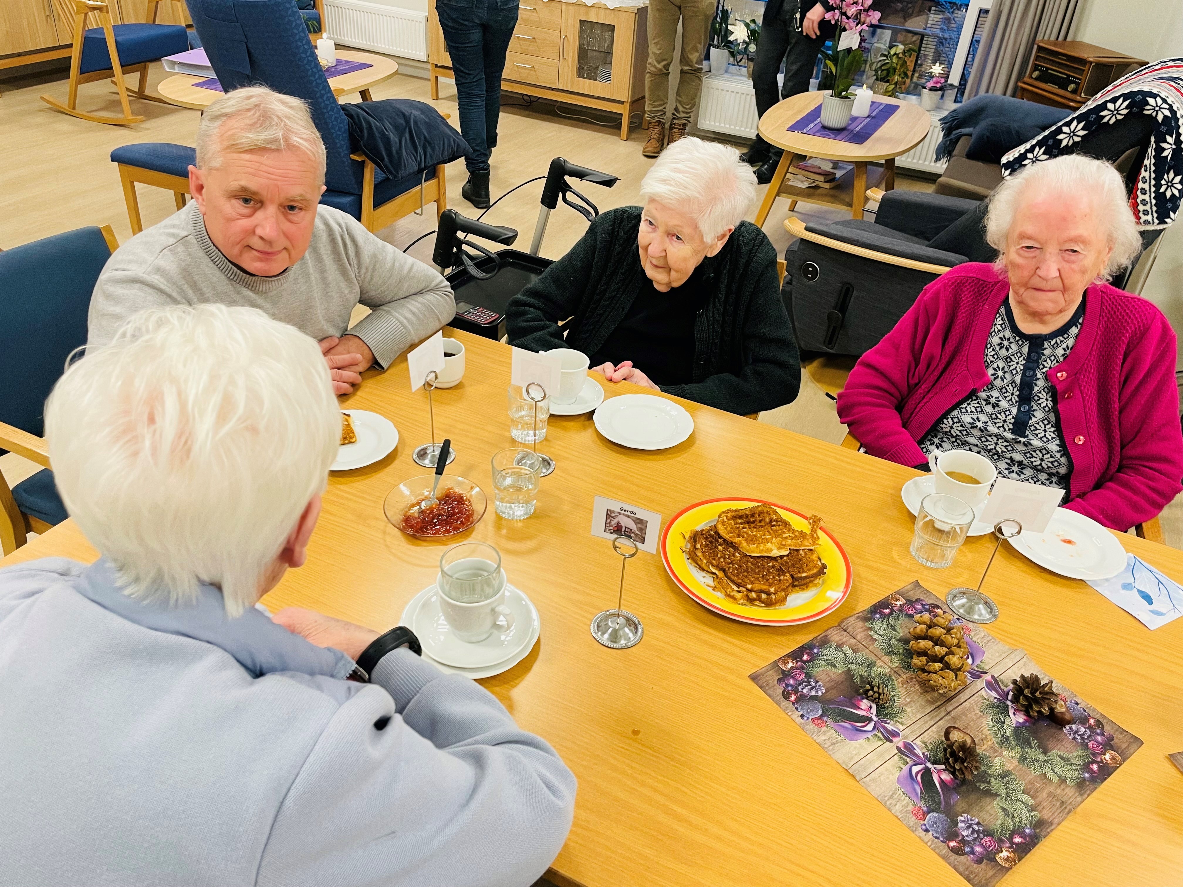 Mann i samtale med eldre mennesker rundt et bord på en institusjon.