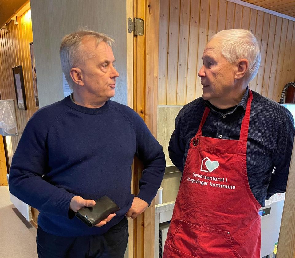 Knut Storberget står i døra til kjøkkenet på seniorsenteret sammen med eldre frivillig mann i rødt forkle,